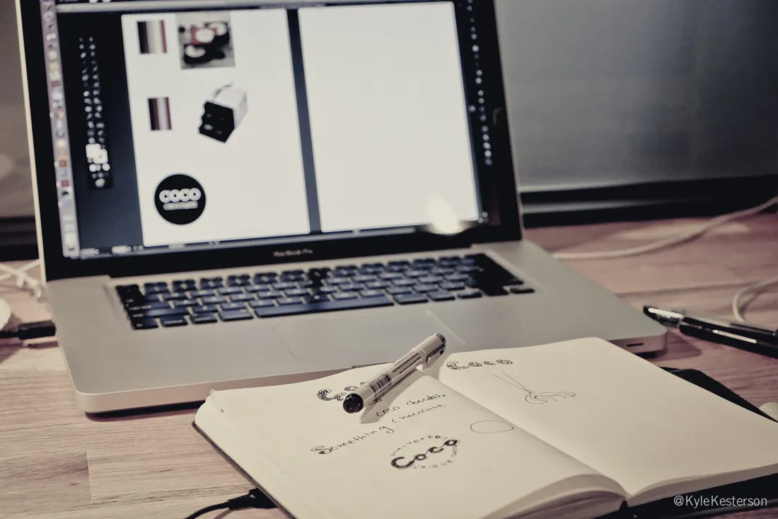 A laptop with brand comps beside an open sketchbook of hand-lettered logo studies