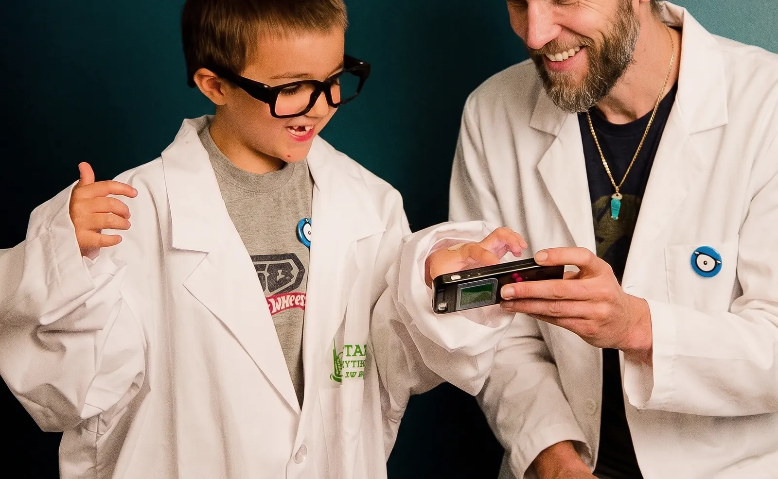 Kyle and a boy in lab coats, both grinning at a handheld device
